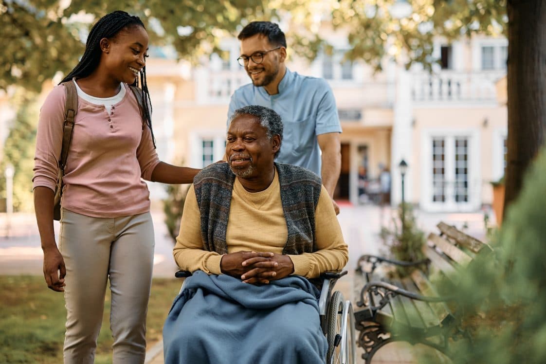 A carer pushing an older person in a wheelchair while another carer is talking to him.